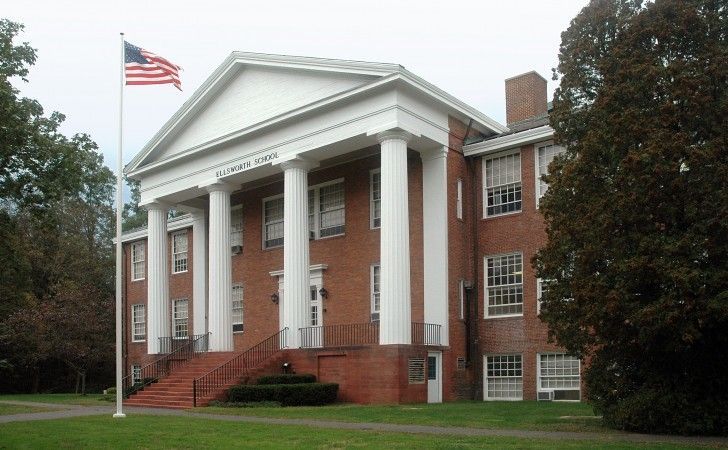 A red brick building with white columns and a triangular pediment, featuring an American flag on a pole to the left.