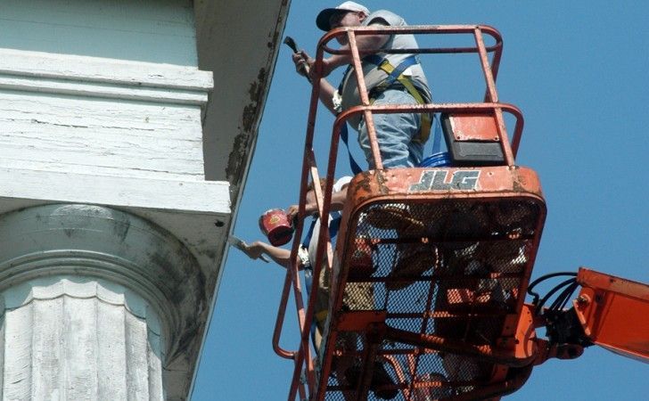 Two people in an orange JLG aerial lift paint the exterior of a building above a large white column against a blue sky.