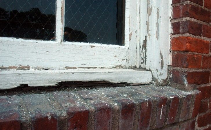 Close-up of a weathered, white-painted wooden window frame with peeling paint and wood rot against a red brick wall.