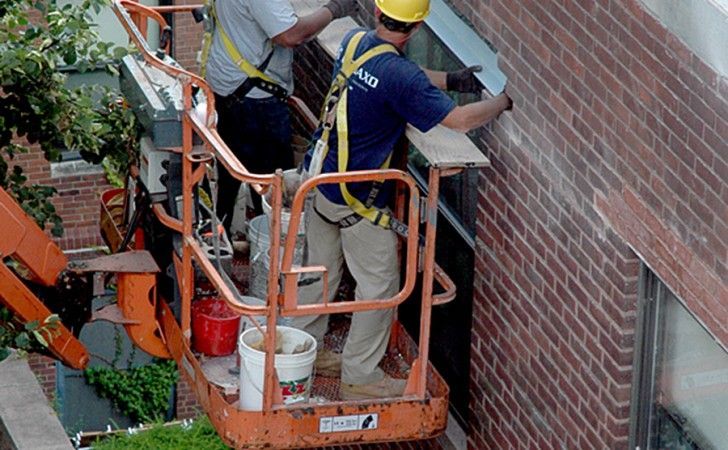 Two workers in safety harnesses stand in an orange aerial lift, installing a metal piece on a brick building exterior.