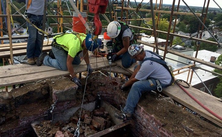 Construction workers in safety gear operate a crane to lift debris from a rooftop repair site surrounded by scaffolding.