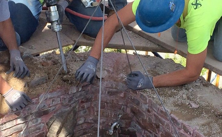Workers wearing hard hats and gloves use tools to remove bricks from a circular construction site.