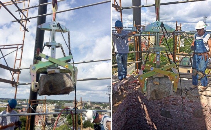 Two construction workers on high scaffolding use a mechanical lifting clamp to move a large stone block.