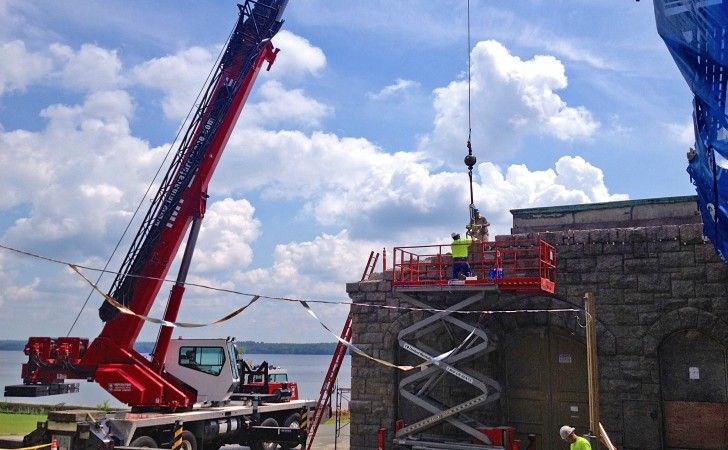 A red crane lifts a load to workers on a red scissor lift next to a historic stone building by the water.