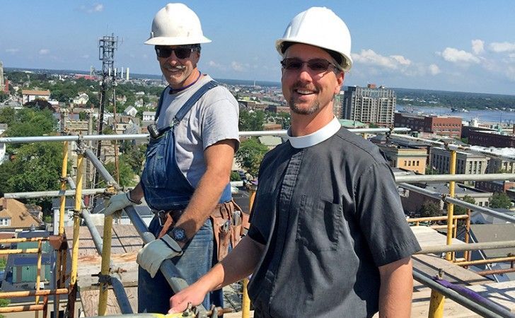 Two people in hard hats and work attire stand on scaffolding overlooking a city.