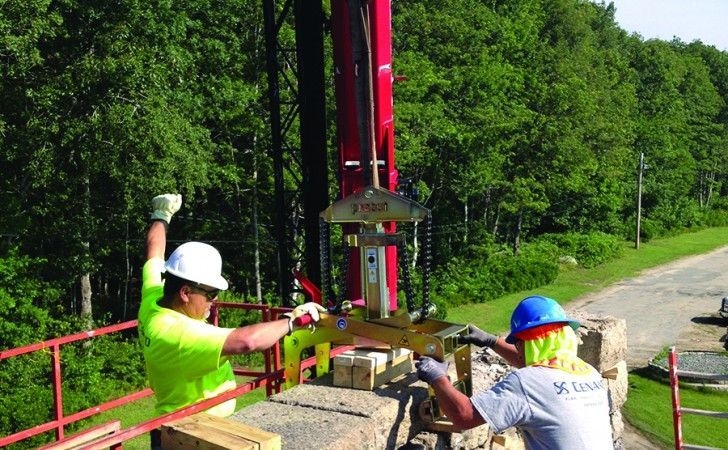 Two construction workers in safety gear operate a yellow stone-lifting clamp on a wall at a worksite.