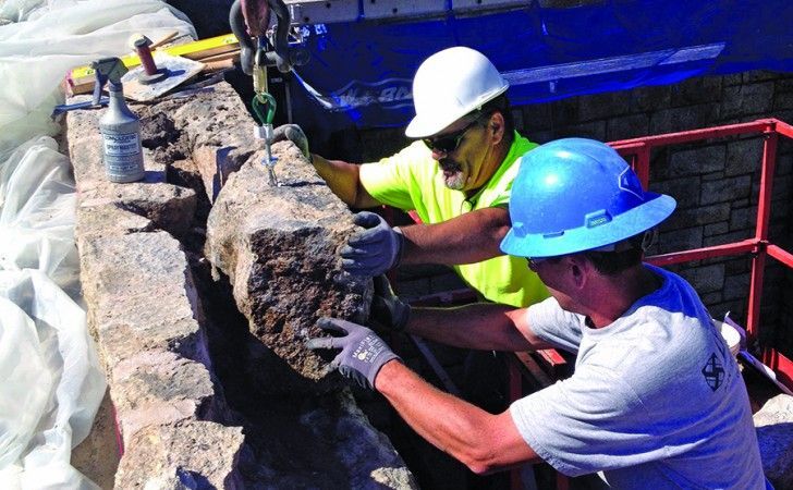 Two workers in hard hats use a crane to position a large, rough stone block into a masonry wall on a construction site.