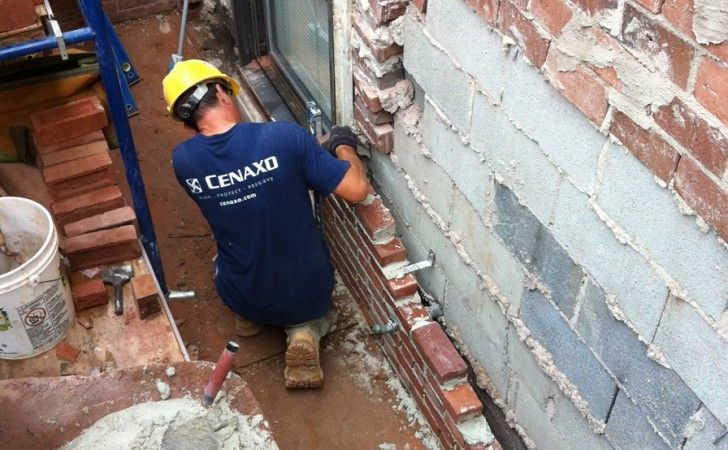 A construction worker in a yellow hard hat and blue shirt lays bricks along the base of a building wall.