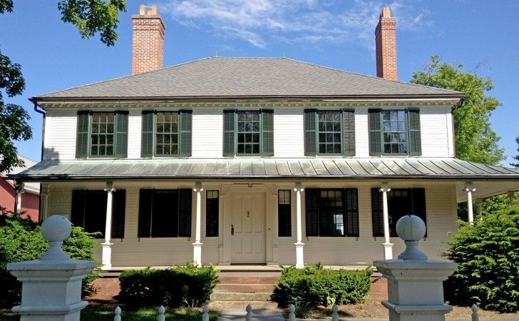A two-story white house with dark shutters, a front porch with pillars, and two brick chimneys under a blue sky.