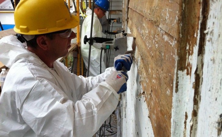 Two workers in white protective suits and hard hats strip old material from a wooden wall on a construction site.