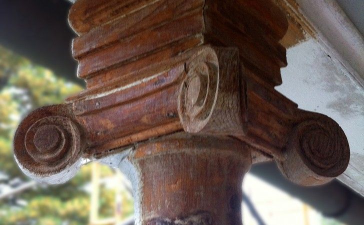A close-up of a weathered, dark brown wooden column capital featuring distinctive carved spiral volutes.