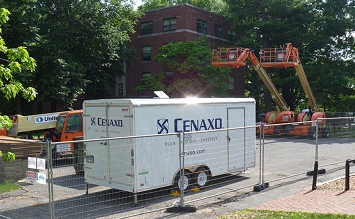 A white Cenaxo trailer sits in an outdoor area near construction equipment, including orange aerial lifts and fencing.