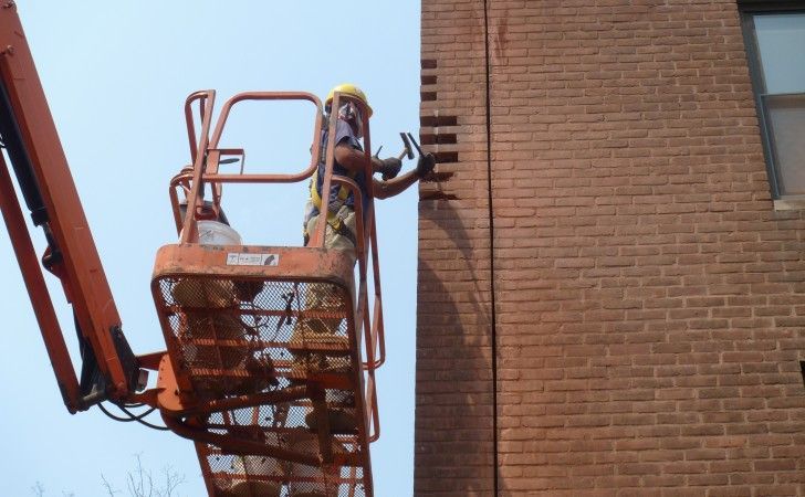 A person in a hard hat and safety harness works on a brick wall from a raised orange aerial lift.