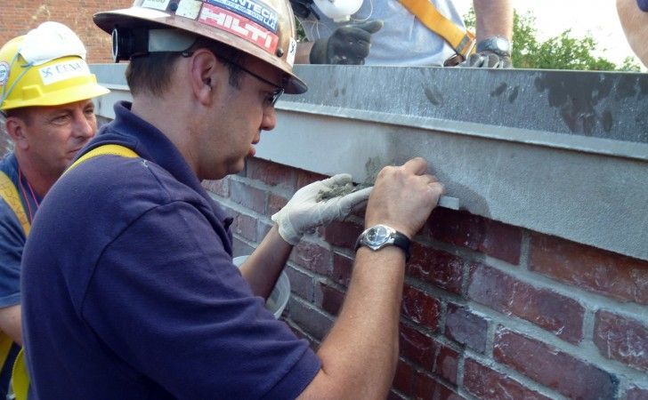A construction worker applies mortar to a brick wall under a stone cap while a colleague observes nearby.