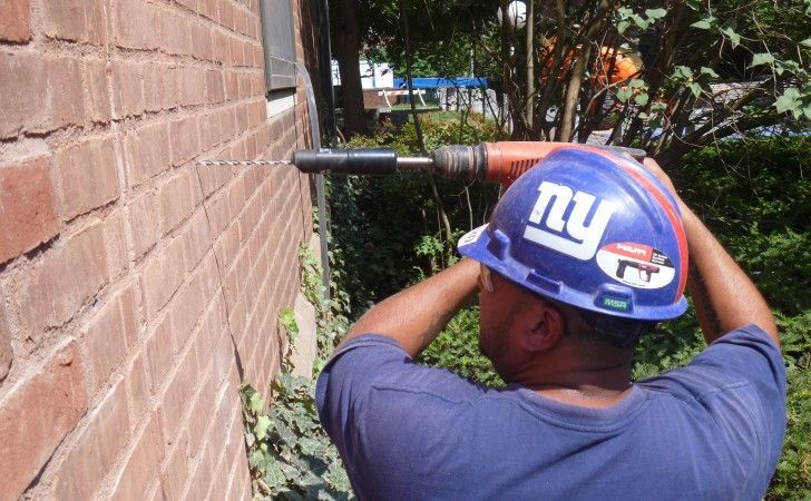 A worker wearing a blue New York Giants hard hat uses a power drill to bore a hole into an exterior brick wall.