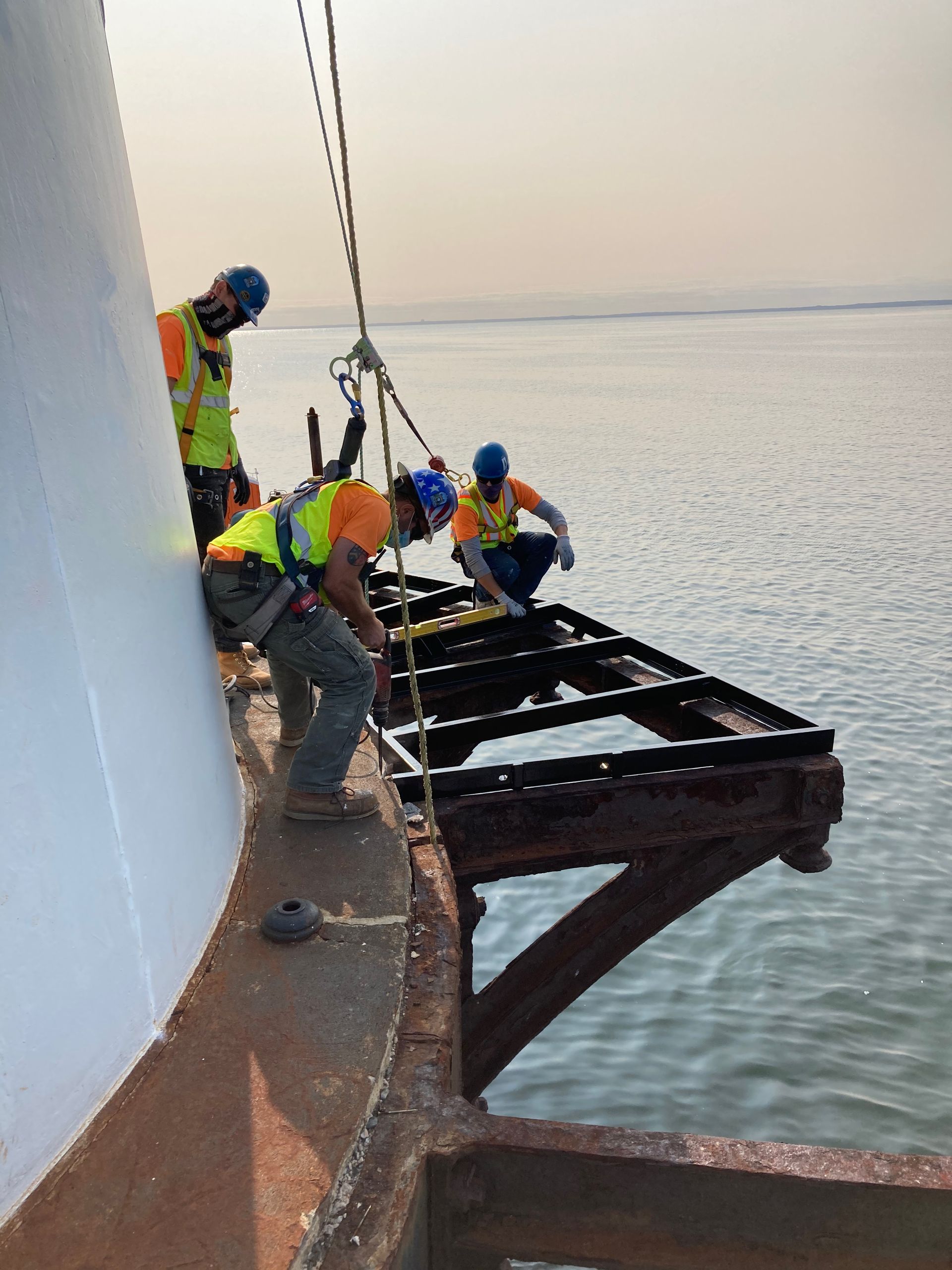 Three workers in safety gear and hard hats perform maintenance on a metal platform attached to a white pillar over water.