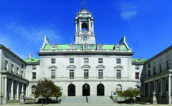 A stately light-gray stone building with a green roof, a central clock tower, and pillared wings under a clear blue sky.