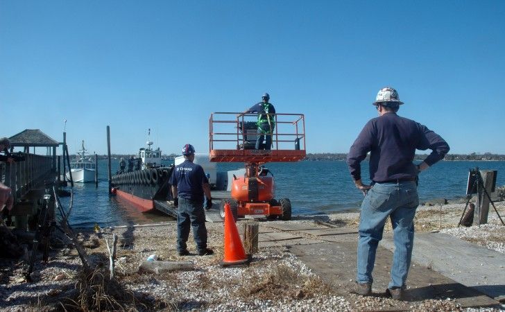 Three workers oversee a scissor lift operating near a boat ramp and a vessel on a sunny day at the waterfront.