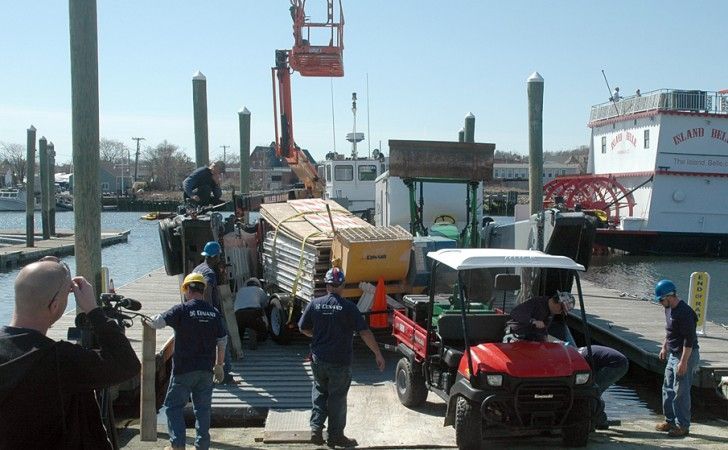 Workers on a wooden pier load construction materials onto a boat near a dock with a utility vehicle and a lift nearby.
