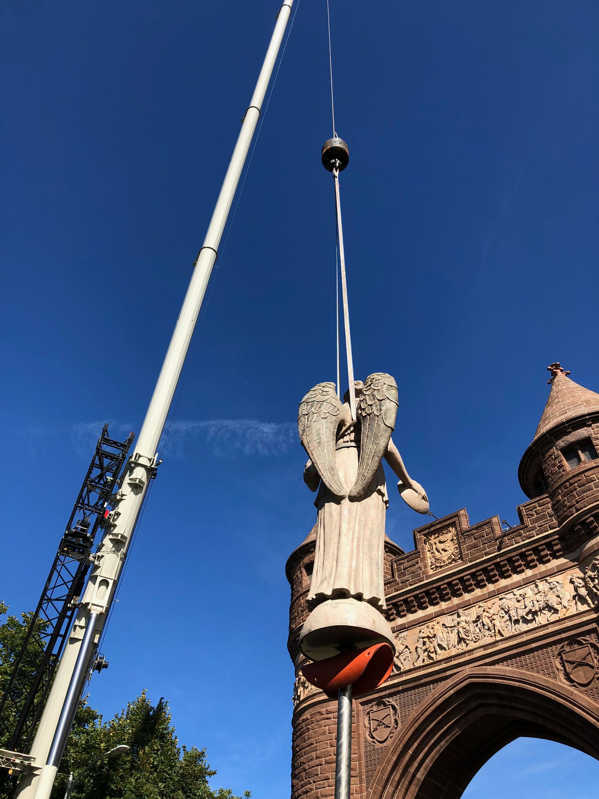 A stone angel statue being hoisted by a crane in front of a historic stone archway under a clear blue sky.