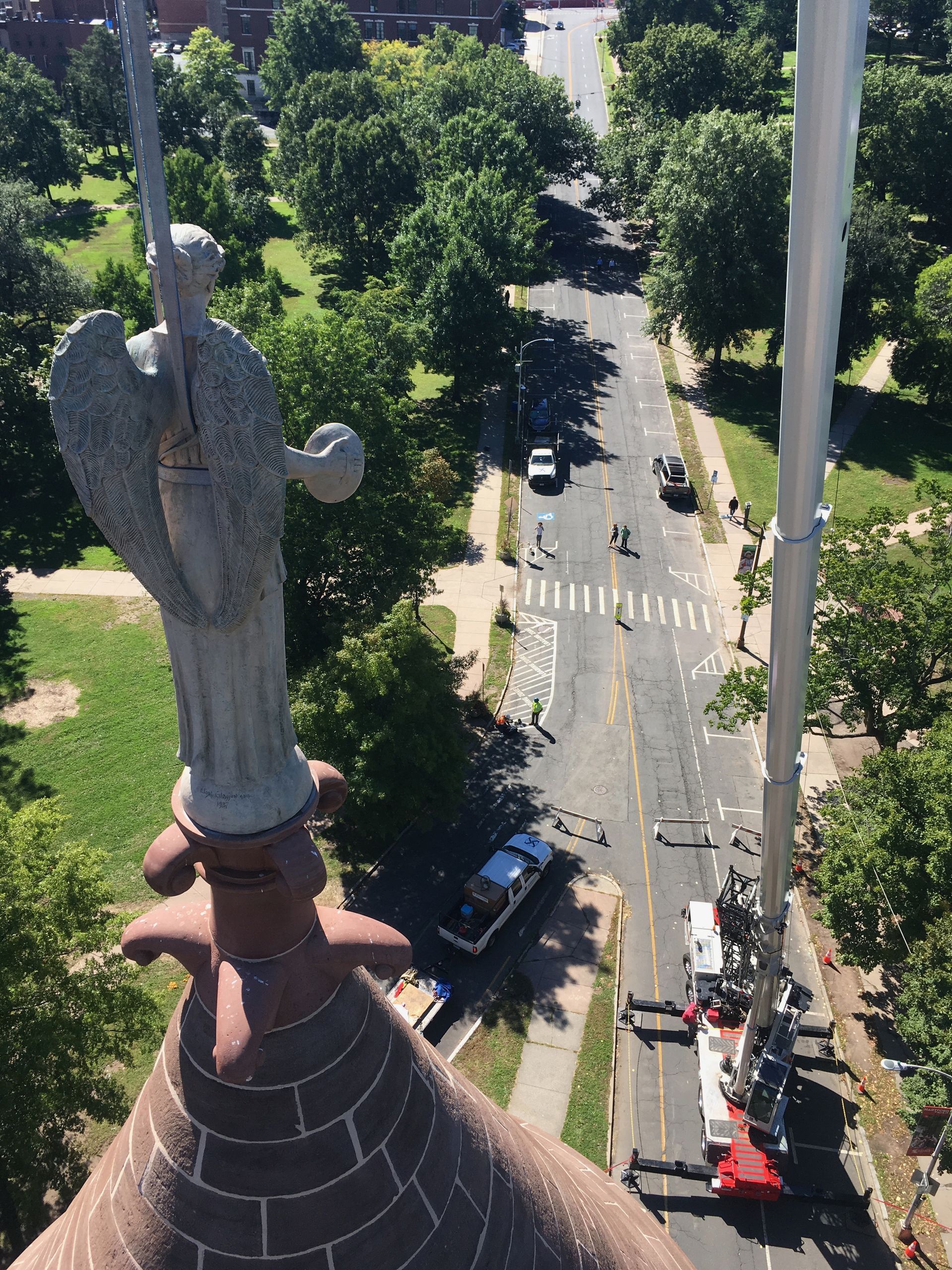 A high-angle view of a stone angel statue atop a church spire, with a large construction crane parked on the road below.