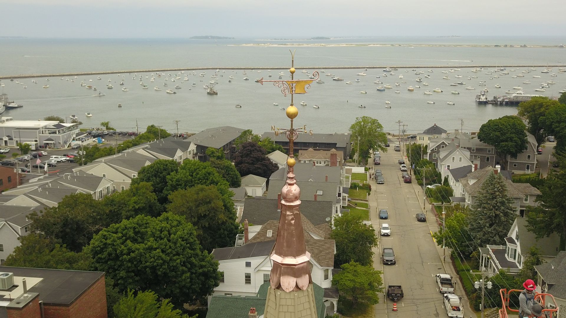 An elevated view of a coastal town street leading to a harbor filled with boats, featuring a copper church steeple.