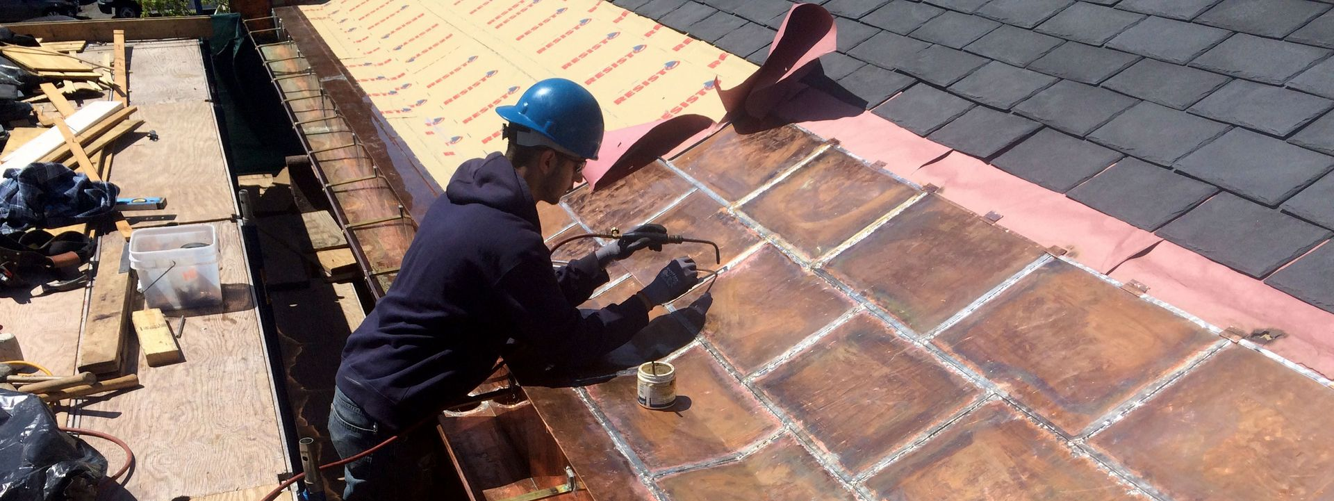 A worker in a blue hard hat installs metal roofing panels on a residential roof.