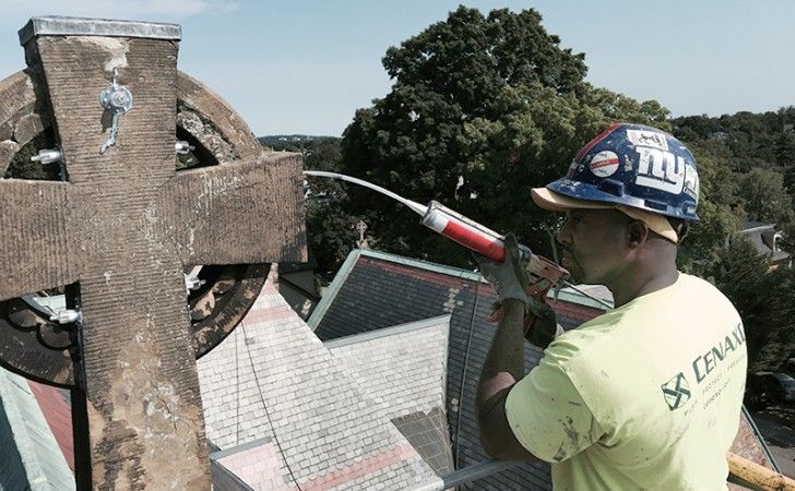 A construction worker wearing a hard hat uses a caulk gun to repair a stone cross on a roof.