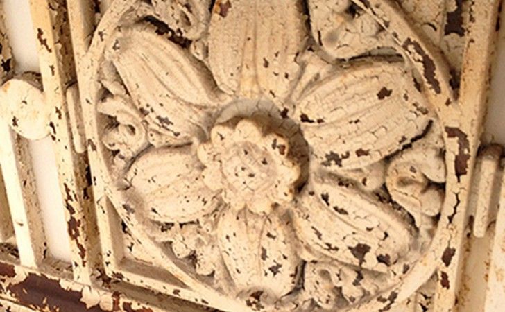 A close-up of a weathered, cream-colored floral relief carving on a decorative metal gate with chipping paint.