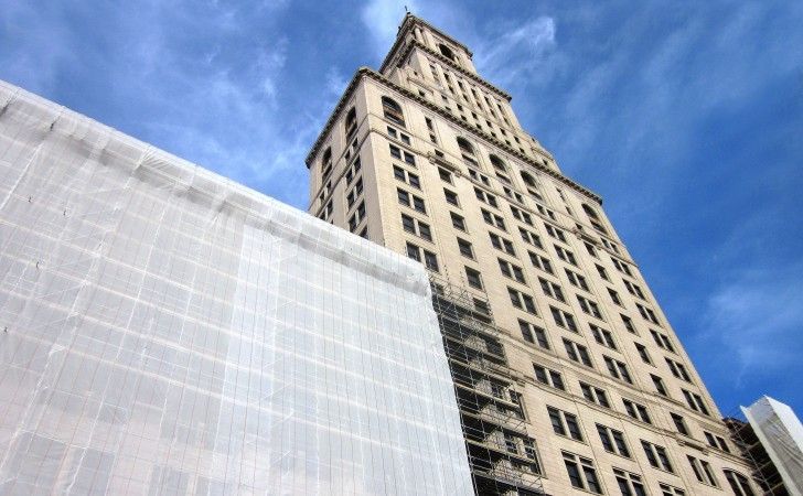 A tall, light-colored historic skyscraper with a tiered tower against a bright blue sky, partially covered by scaffolding.