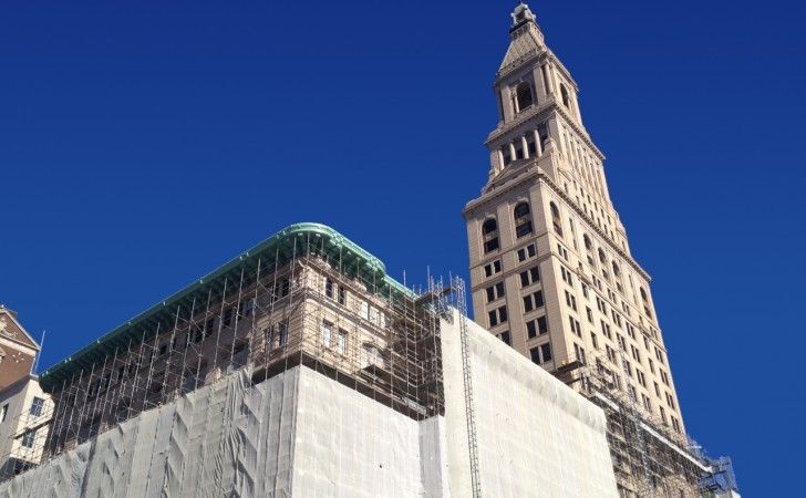 A tall, tan stone clock tower stands beside a building covered in scaffolding and white protective netting.