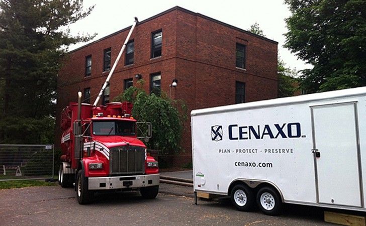 A red construction truck with an extended boom parked next to a white trailer branded with the Cenaxo logo near a building.