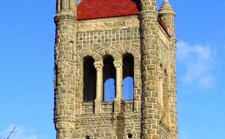 A stone church tower with three arched belfry windows and a red roof against a blue sky.