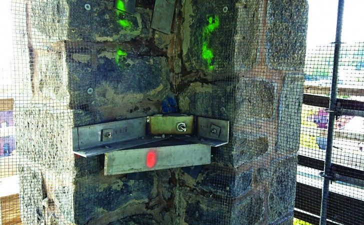 A corner of a stone column showing structural steel reinforcement, spray-painted markings, and wire mesh on a work site.