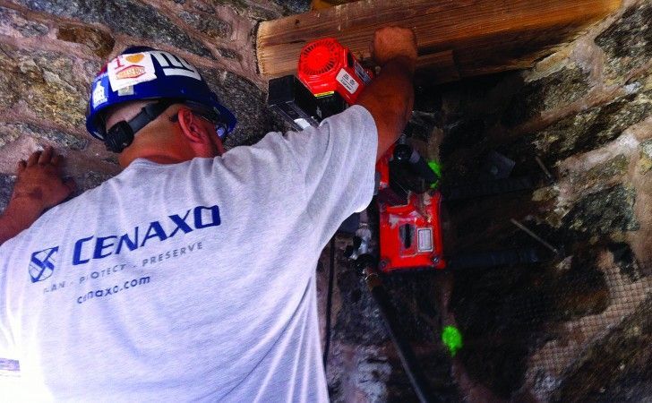 A construction worker in a hard hat uses a power tool to work on a stone structure.