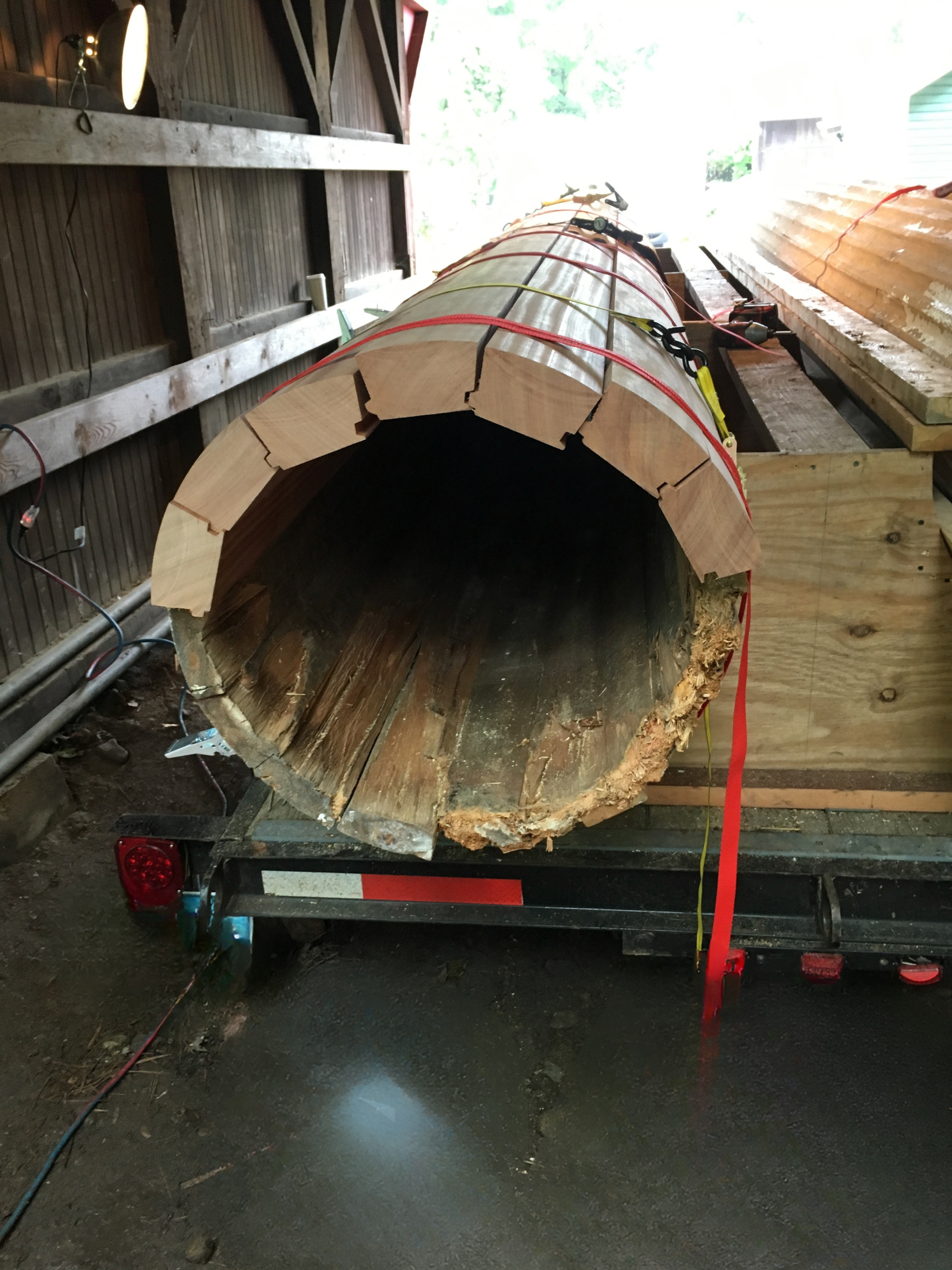 A large, hollowed-out wooden column section sits on a flatbed trailer in a barn-like structure.