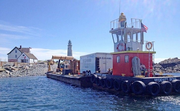 A tugboat pushing a barge with a shipping container and construction equipment, near a lighthouse and a coastal house.