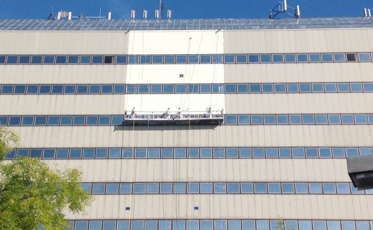 Workers on a suspended platform perform maintenance on the exterior of a light-colored, multi-story office building.