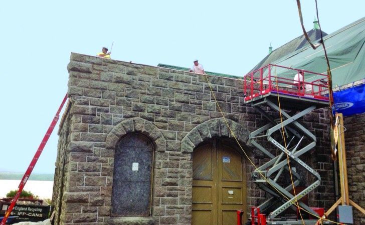 Workers in hard hats on the roof and a scissor lift perform maintenance on a stone building with arched doorways.