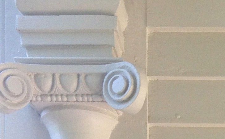 A close-up of a white painted column capital with scroll-like volutes, attached to a light grey brick wall.