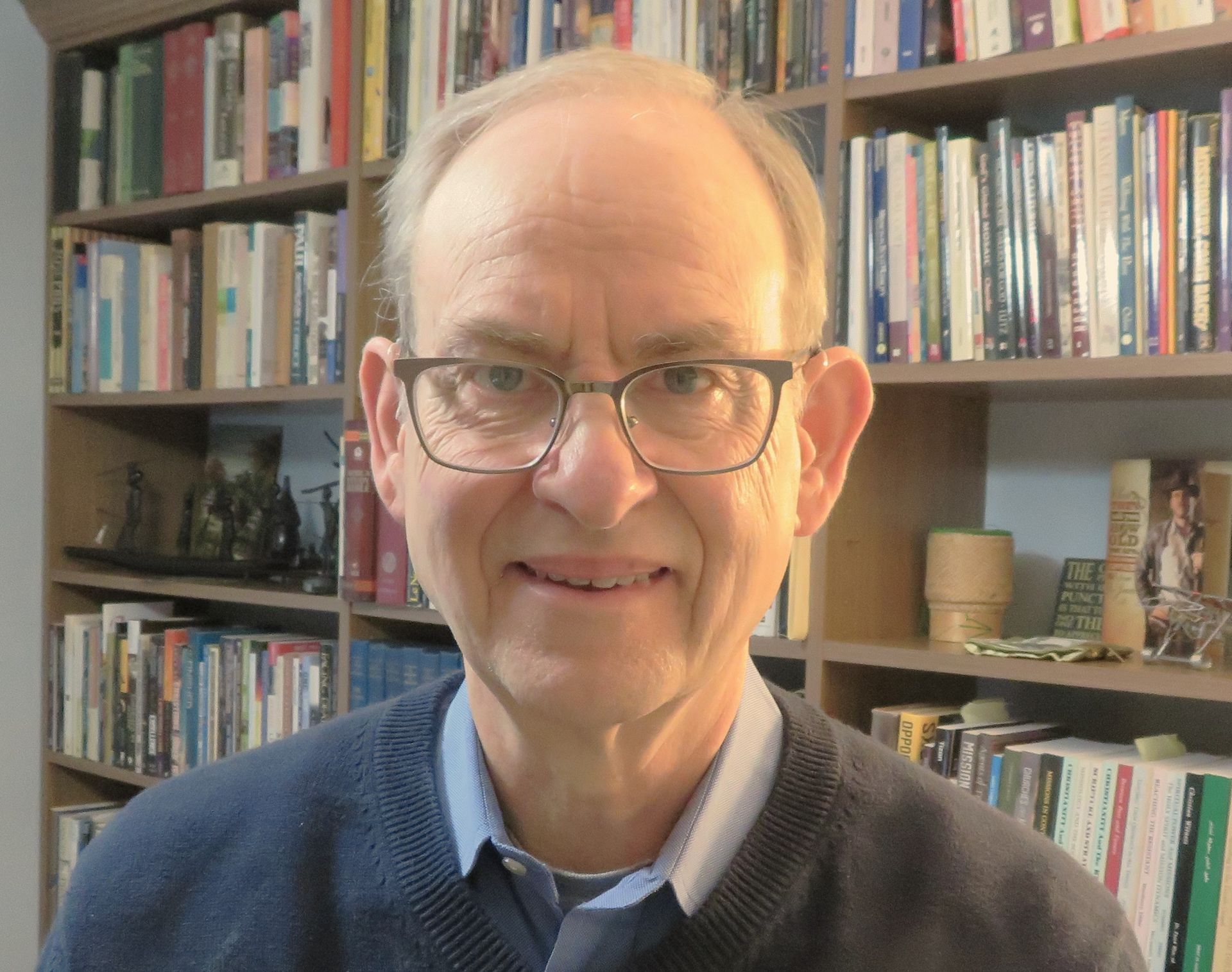 A man wearing glasses is smiling in front of a bookshelf filled with books.