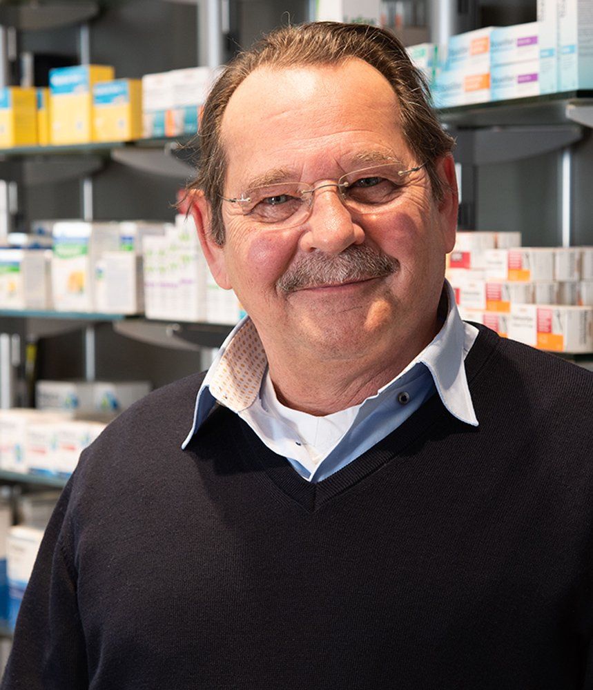 Ein Mann mit Brille und Schnurrbart steht vor einem Apothekenregal.