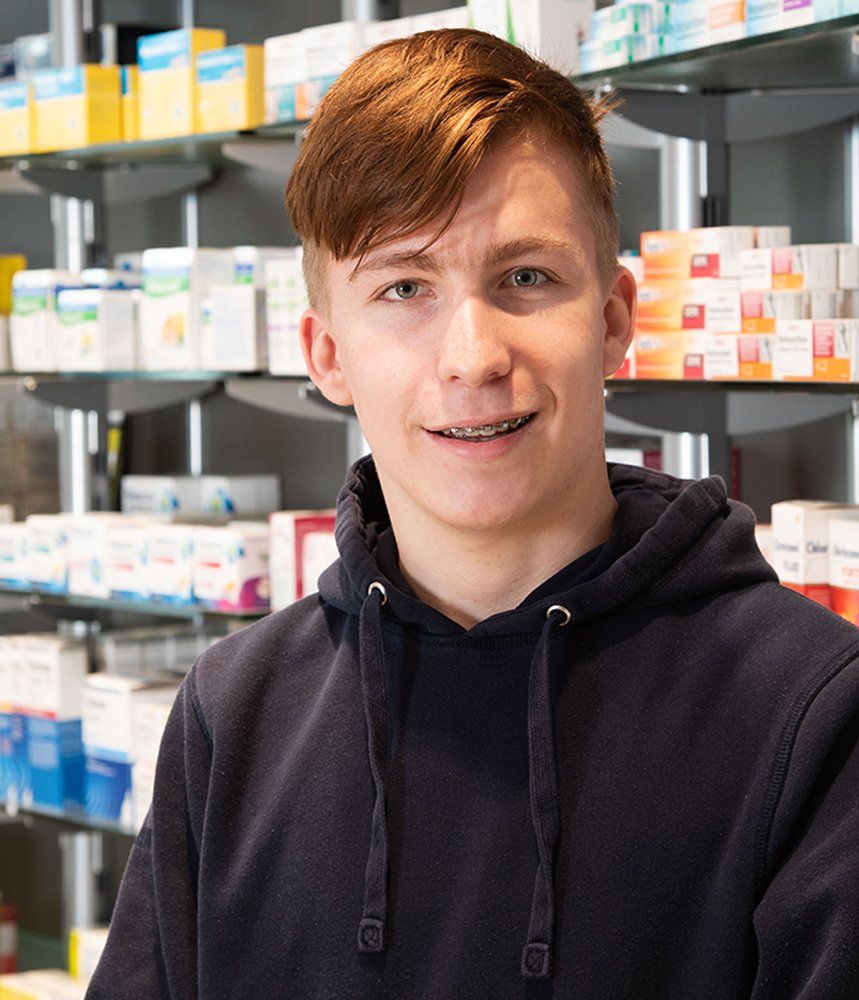 Ein junger Mann mit Kapuzenpullover steht vor einem Apothekenregal.