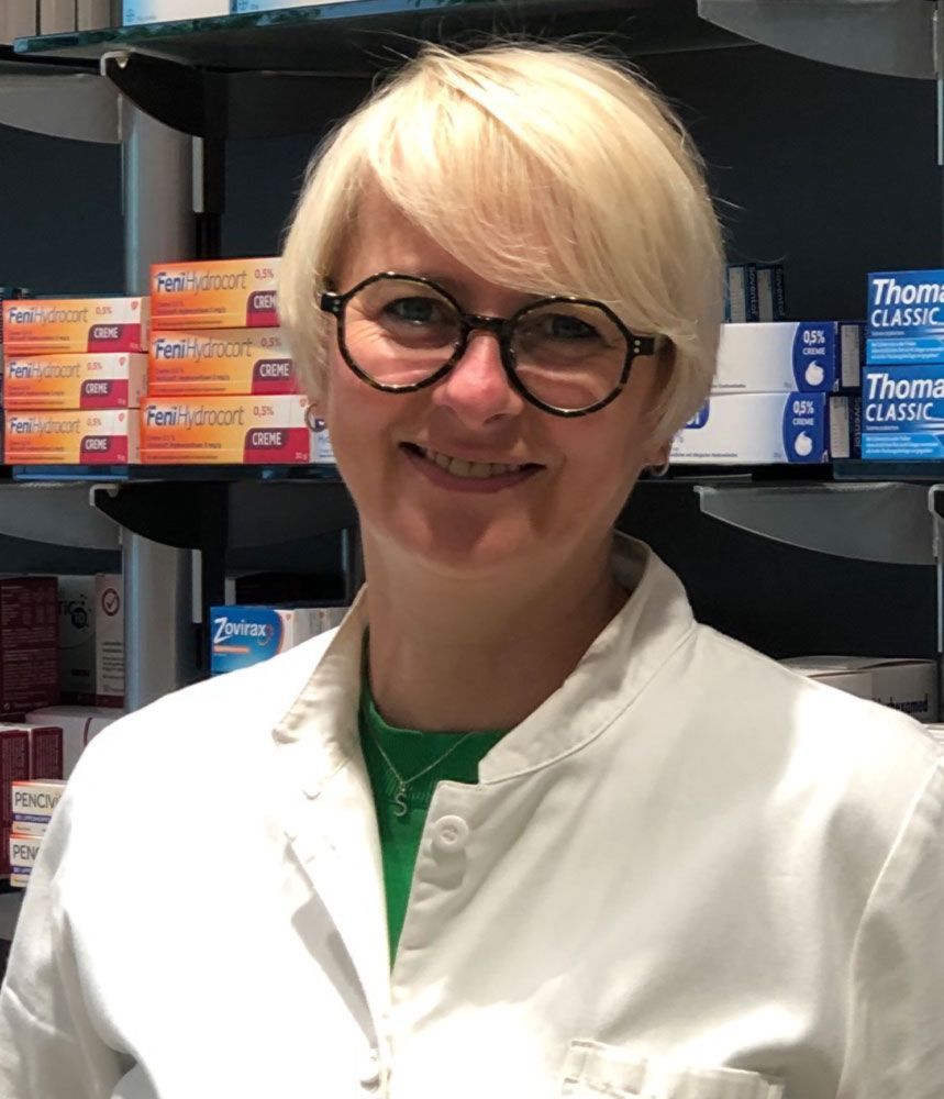 Eine Frau mit Brille und weißem Kittel lächelt in einer Apotheke.