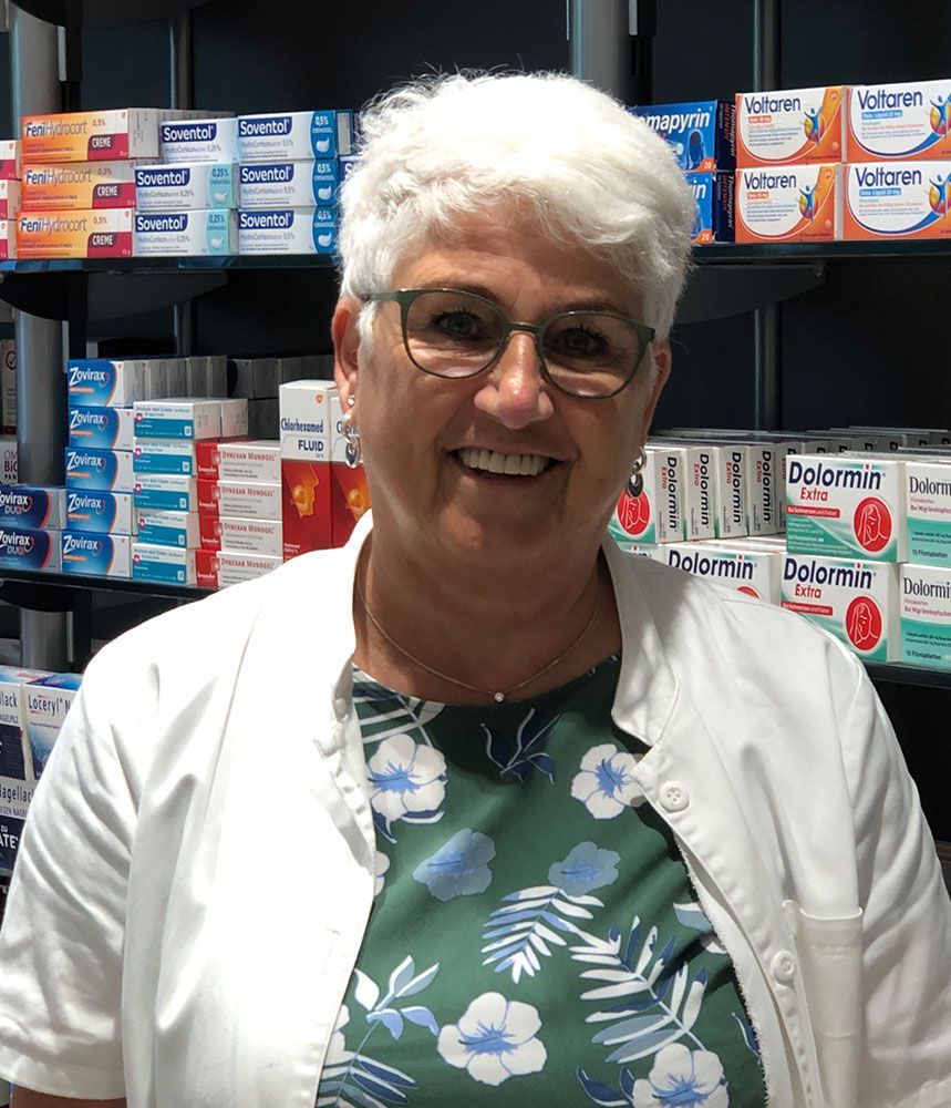 Eine Frau mit Brille und weißem Kittel lächelt in einer Apotheke.