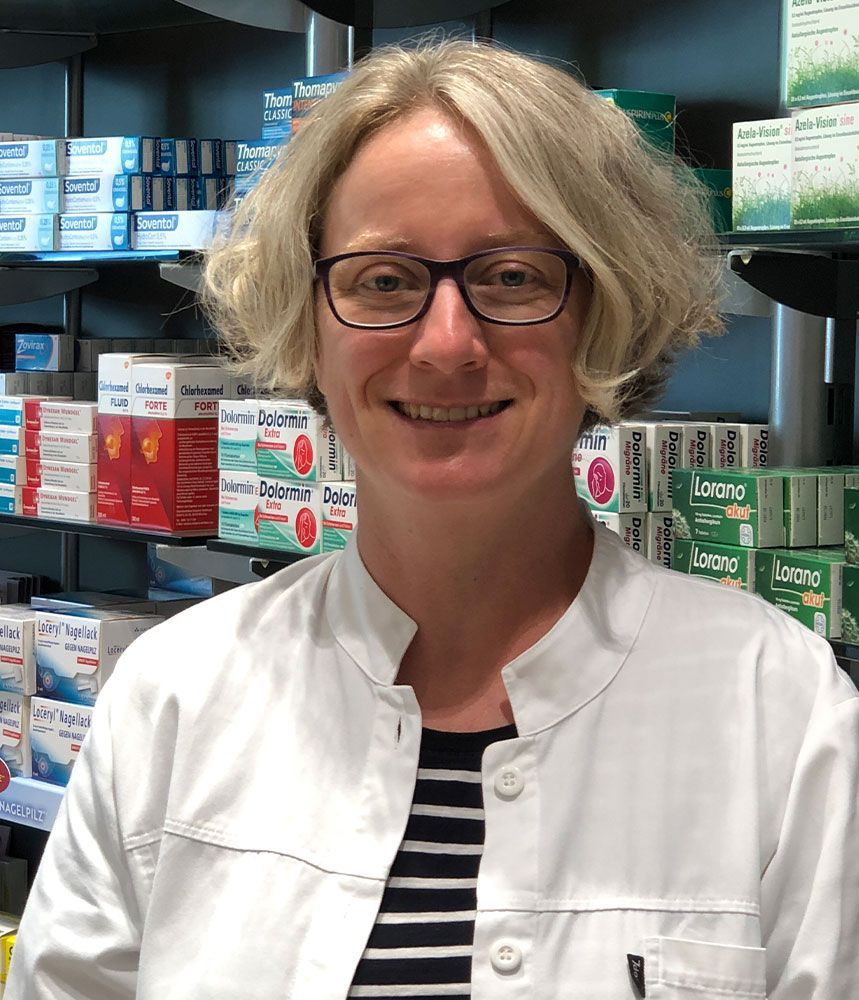 Eine Frau mit Brille und weißem Kittel lächelt in einer Apotheke.