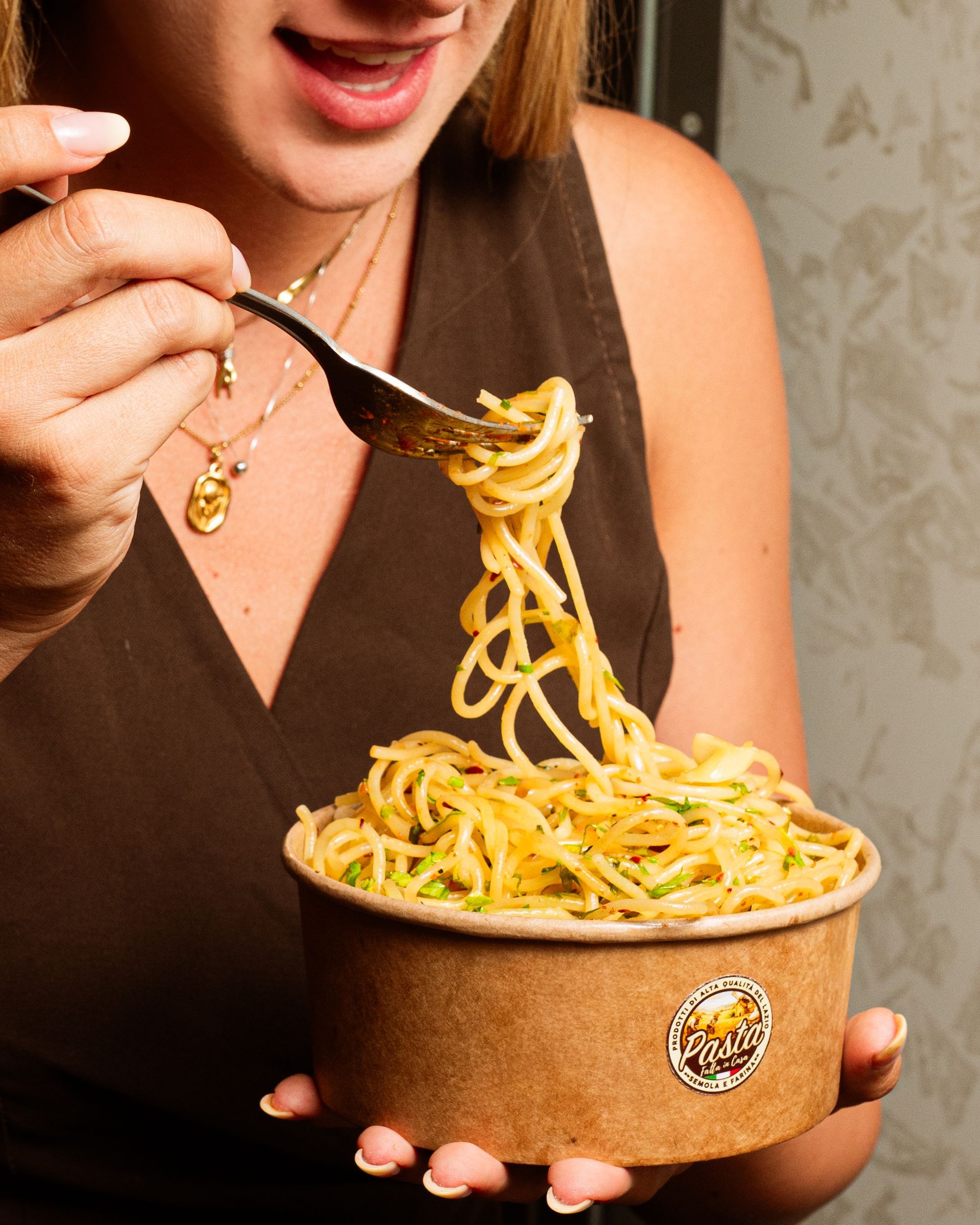 Persona che mangia spaghetti da una ciotola con una forchetta, con indosso una maglietta marrone.