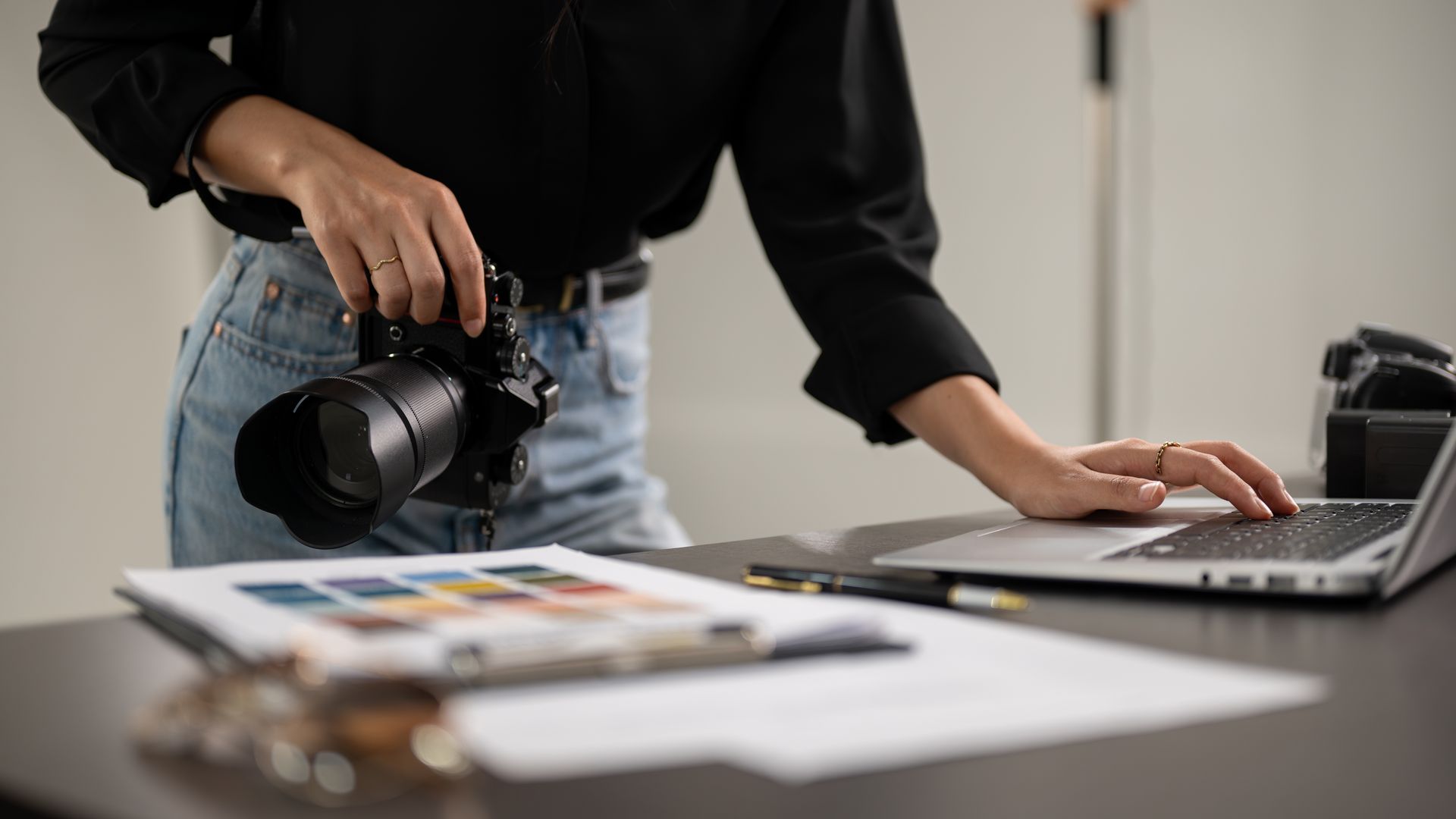 A woman is holding a camera while using a laptop computer.