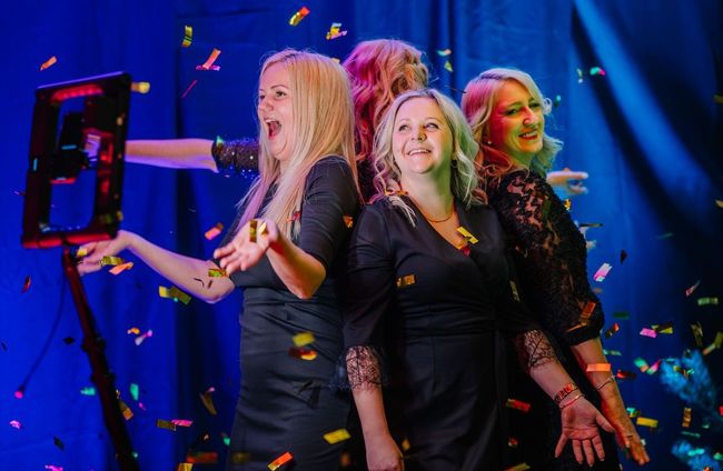 Three women are posing for a picture in a photo booth with confetti falling around them.
