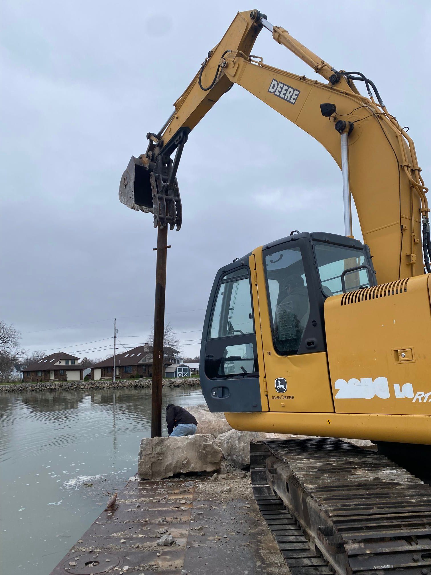 A yellow excavator stands on a shoreline near water, using a vertical pole to work on a concrete block.
